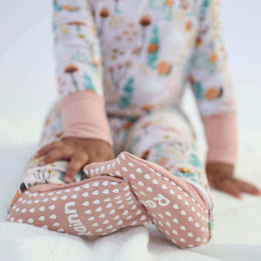 Close-up of a baby's feet in pink organic fabric booties, wearing a floral-patterned onesie, on a soft white blanket.