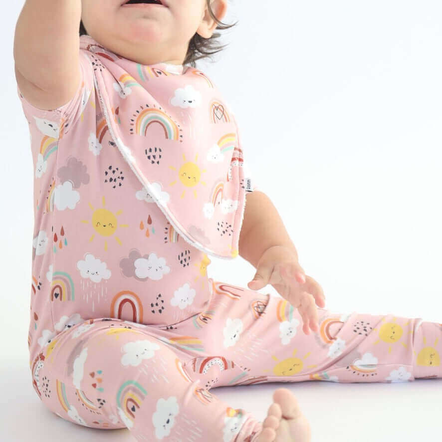 Baby wearing a colorful RainnWeave™ outfit with matching Bibs and Burp Cloths, featuring rainbows and clouds on a pink background.