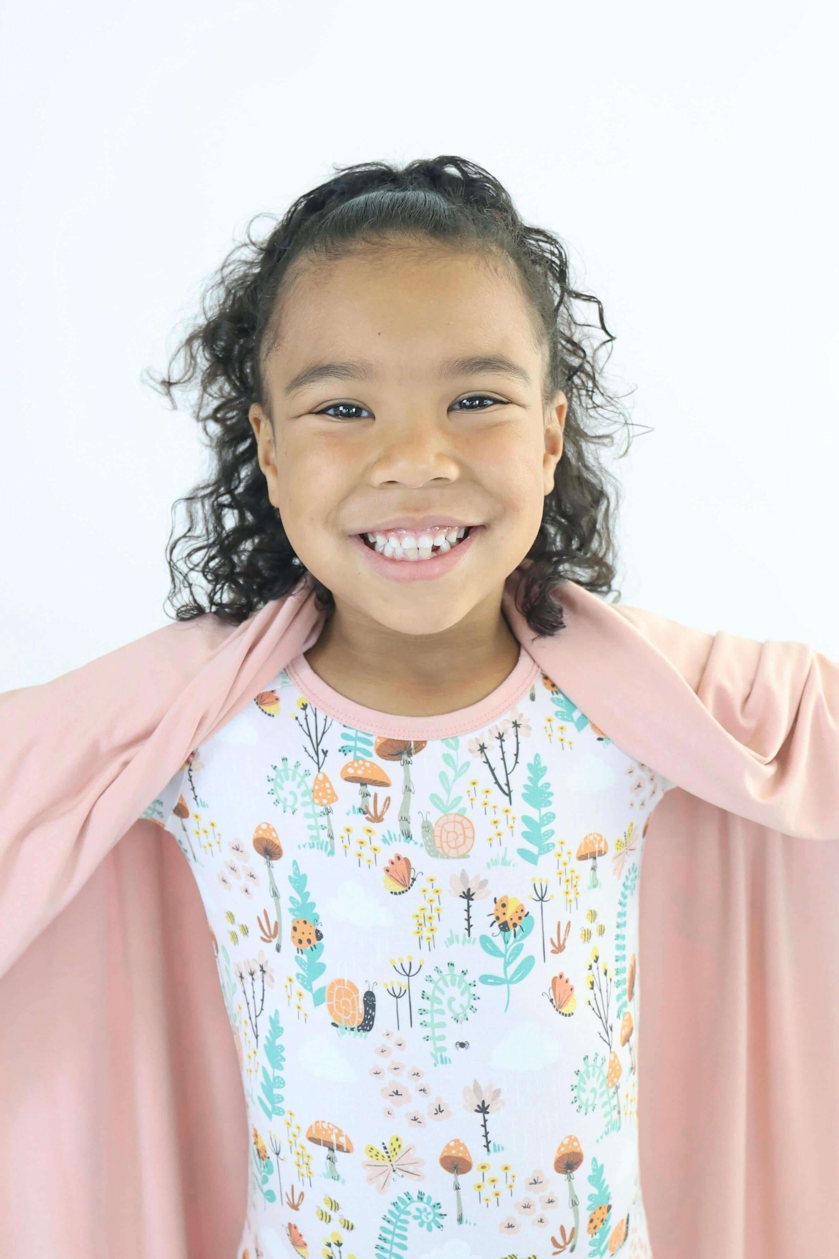 Smiling girl wearing a floral onesie, playfully draped in a soft pink blanket, showcasing a joyful moment.