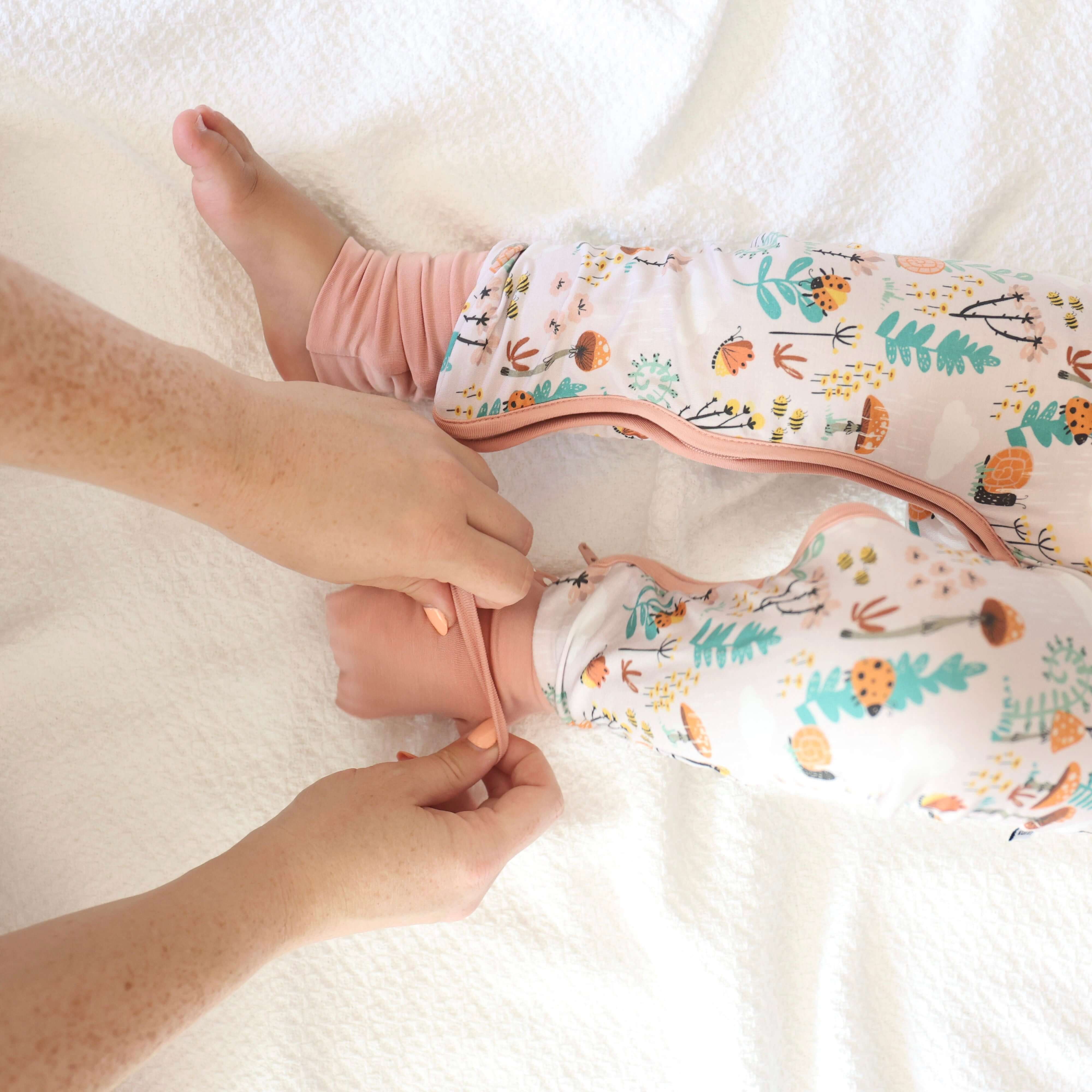 Adult hands zipping up baby in Bamboo Crescent Zipp Jammies with fern and woodland print on white bedding.