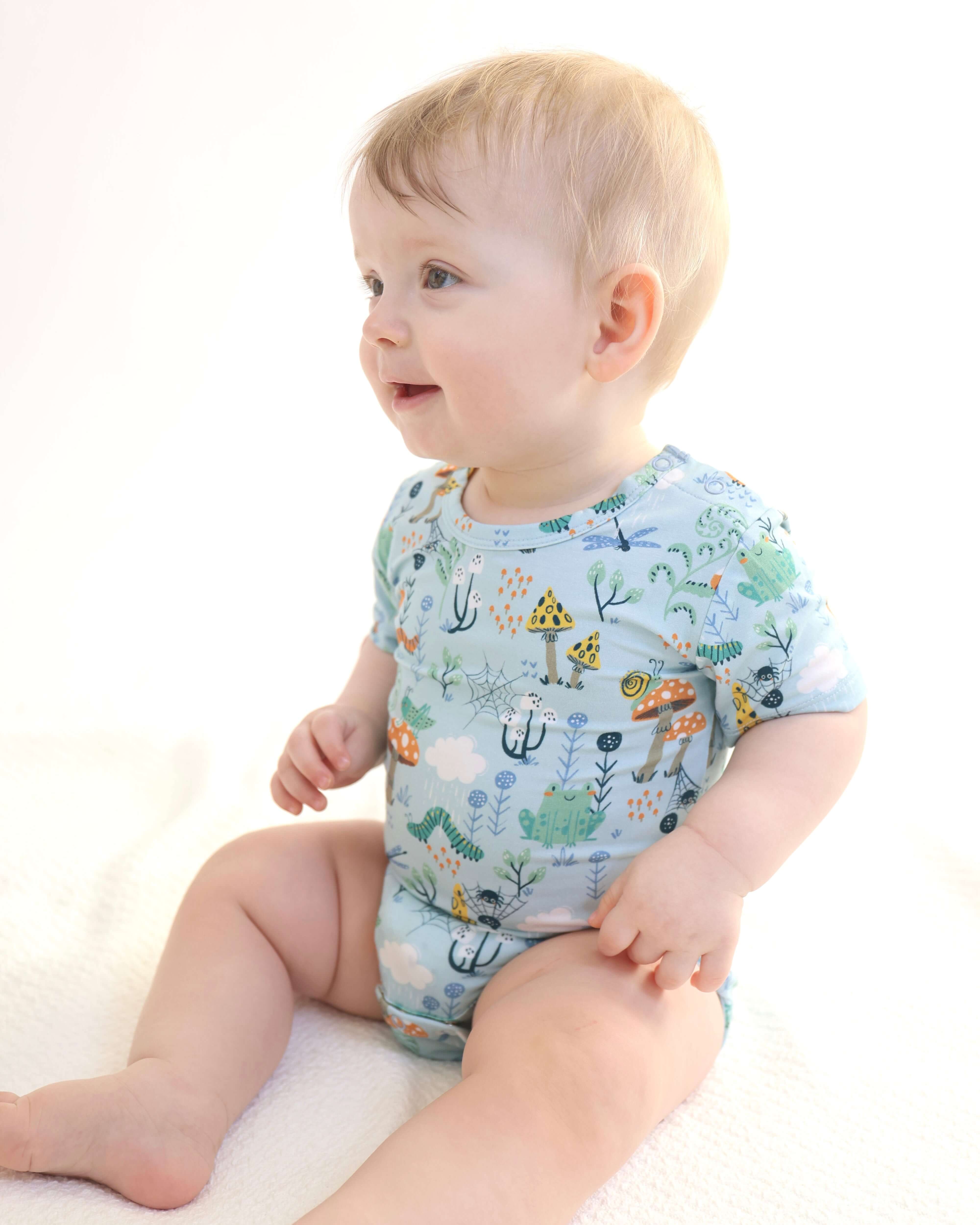Baby wearing a colorful bamboo onesie with nature patterns, smiling and sitting on a soft surface.