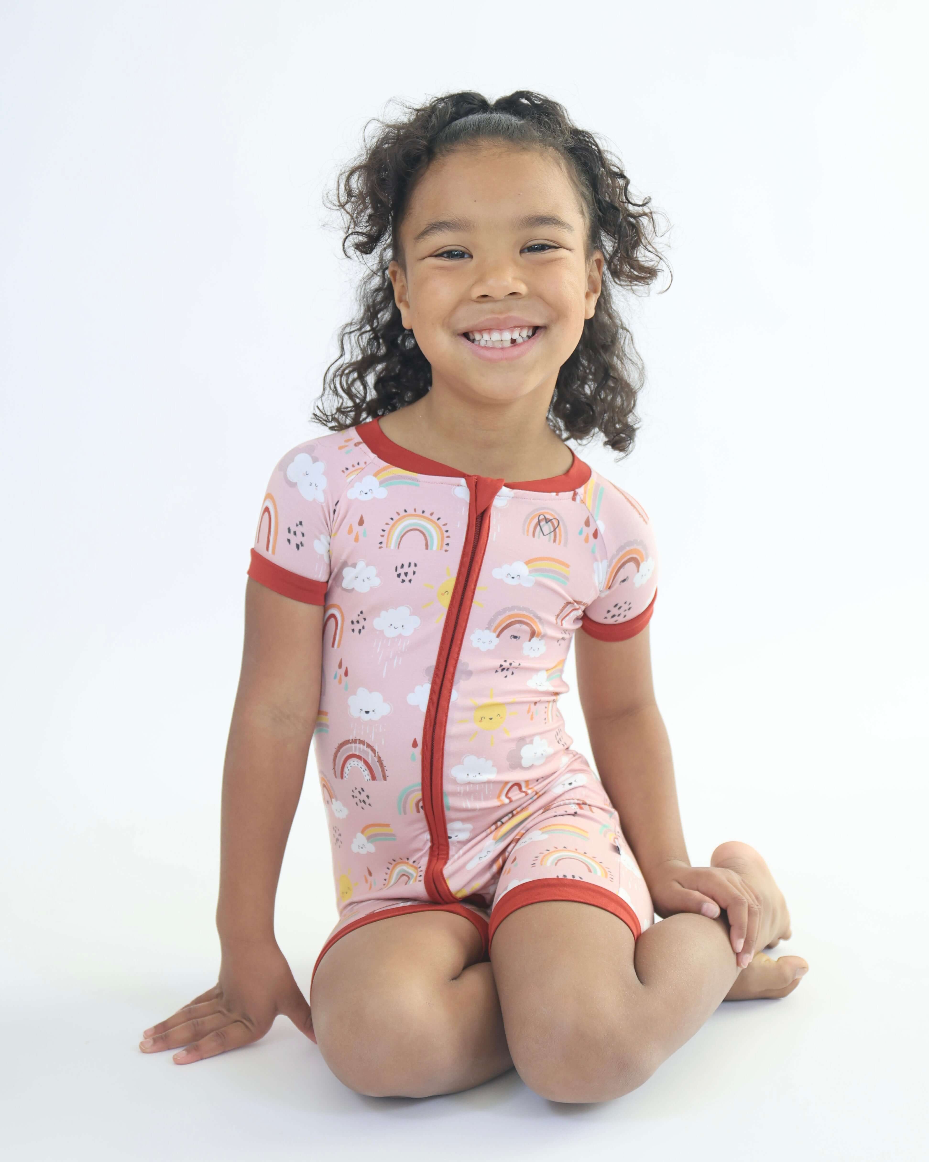 Smiling child in Rainnbow short zip romper with rainbow and cloud print, showcasing breathable and soft bamboo fabric.